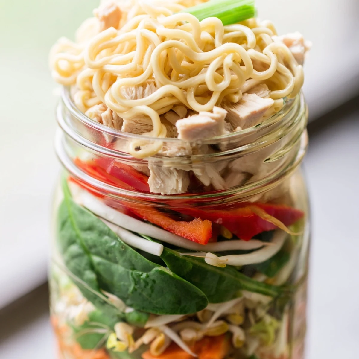 Clear glass mason jars showcase colorful layers of noodles, chicken, and fresh veggies topped with chopped peanuts and cilantro.