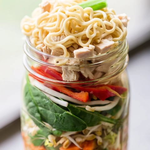 Clear glass mason jars showcase colorful layers of noodles, chicken, and fresh veggies topped with chopped peanuts and cilantro.