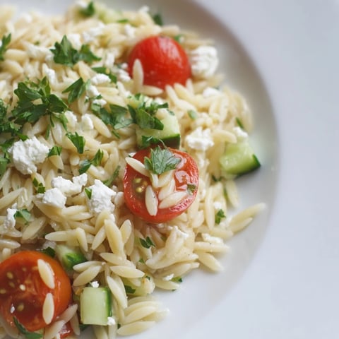 A close-up of Greek Orzo Salad in a white bowl, featuring juicy cherry tomatoes, crisp cucumber, and crumbled feta cheese.