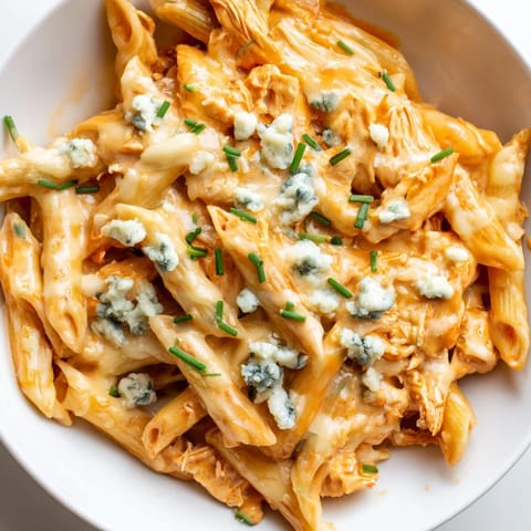 A close-up of Buffalo Chicken Pasta in a white bowl, featuring rotini pasta coated in a rich, cheesy buffalo sauce and garnished with chives.