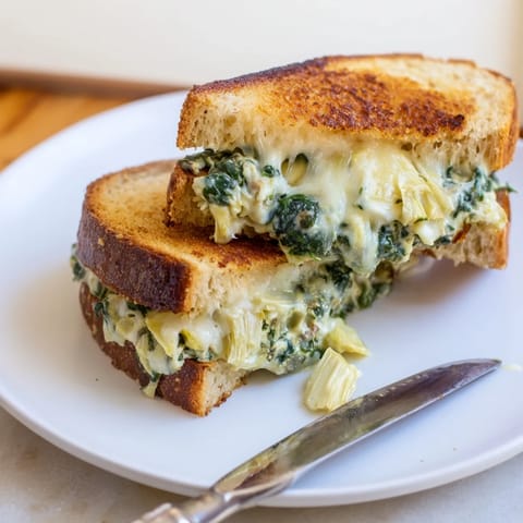 Two halves of a homemade spinach artichoke grilled cheese on a rustic wooden board, melted mozzarella stretching from the crusty bread.  
